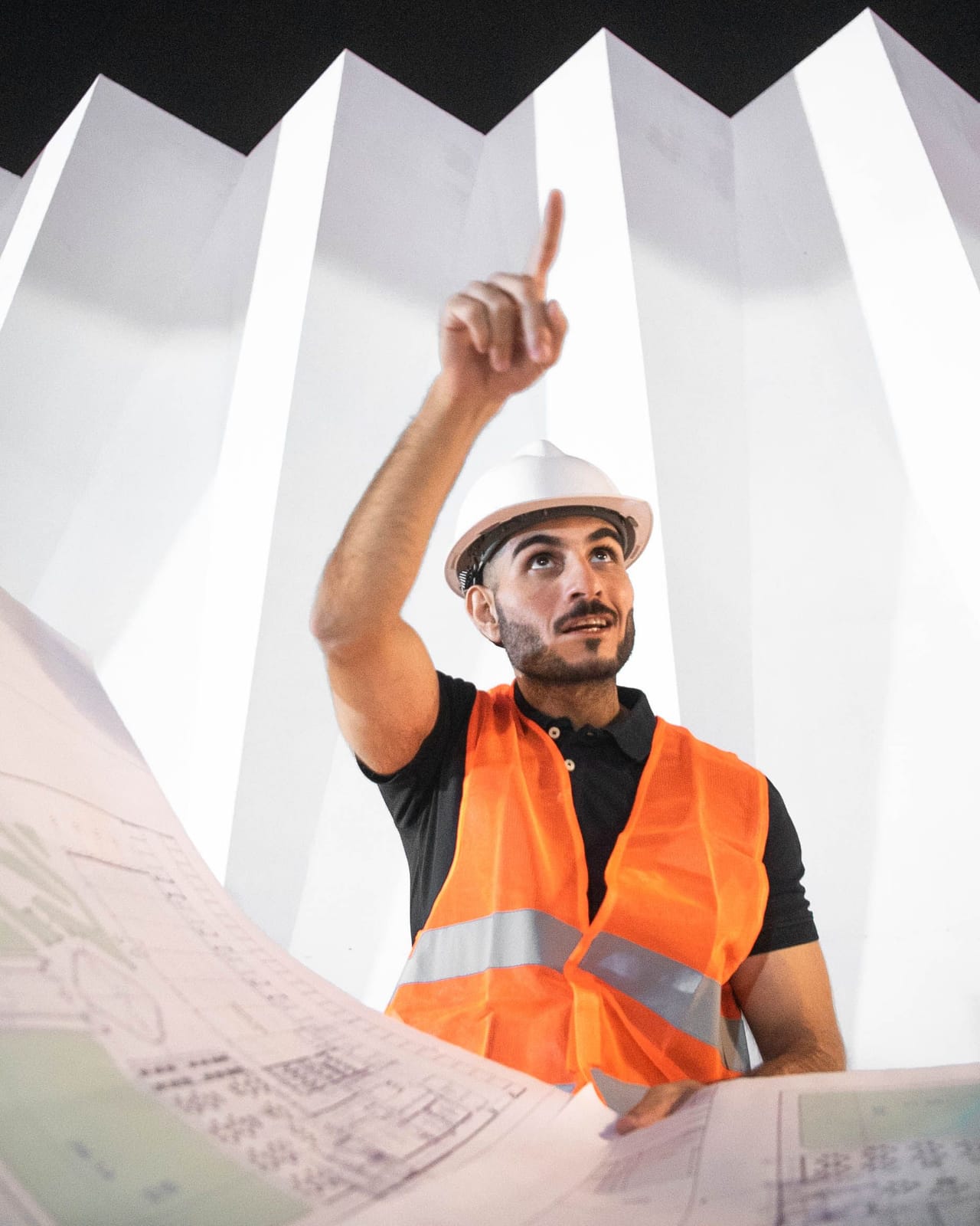 مهندس يرتدي خوذة بيضاء وسترة عاكسة برتقالية يشير إلى الأعلى وأمامه مخططات هندسية